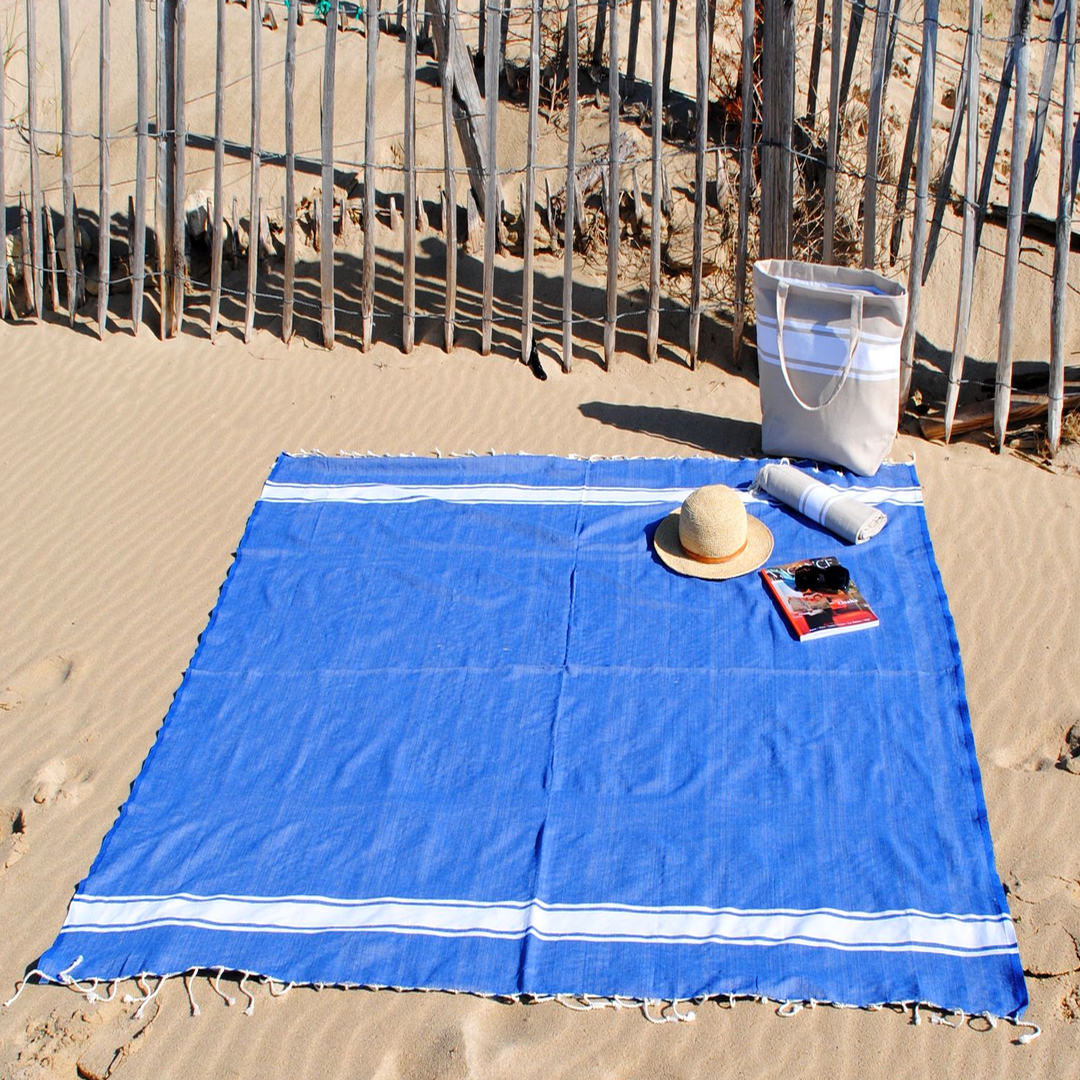 Gran toalla azul real tendida en la arena, sombrero, bolso colocados al lado