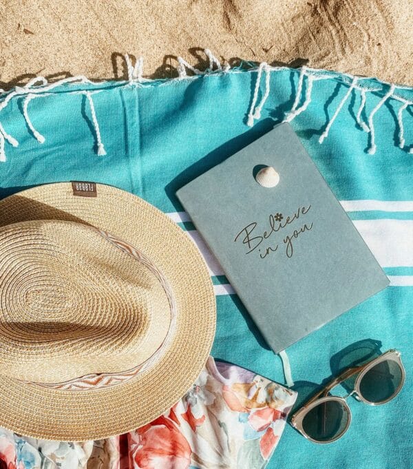 Gran fouta cuadrada azul pavo real con banda blanca sobre fondo arena, con un sombrero, un cuaderno y unas gafas de sol.