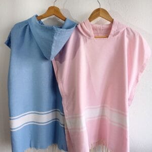 Ponchos con capucha para niños, uno azul cielo y otro rosa, colgados en un perchero.