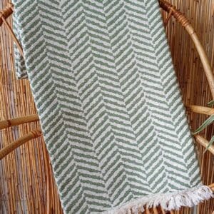 Manta de sofá verde oliva con motivos en espiga colocada sobre un sillón de ratán.